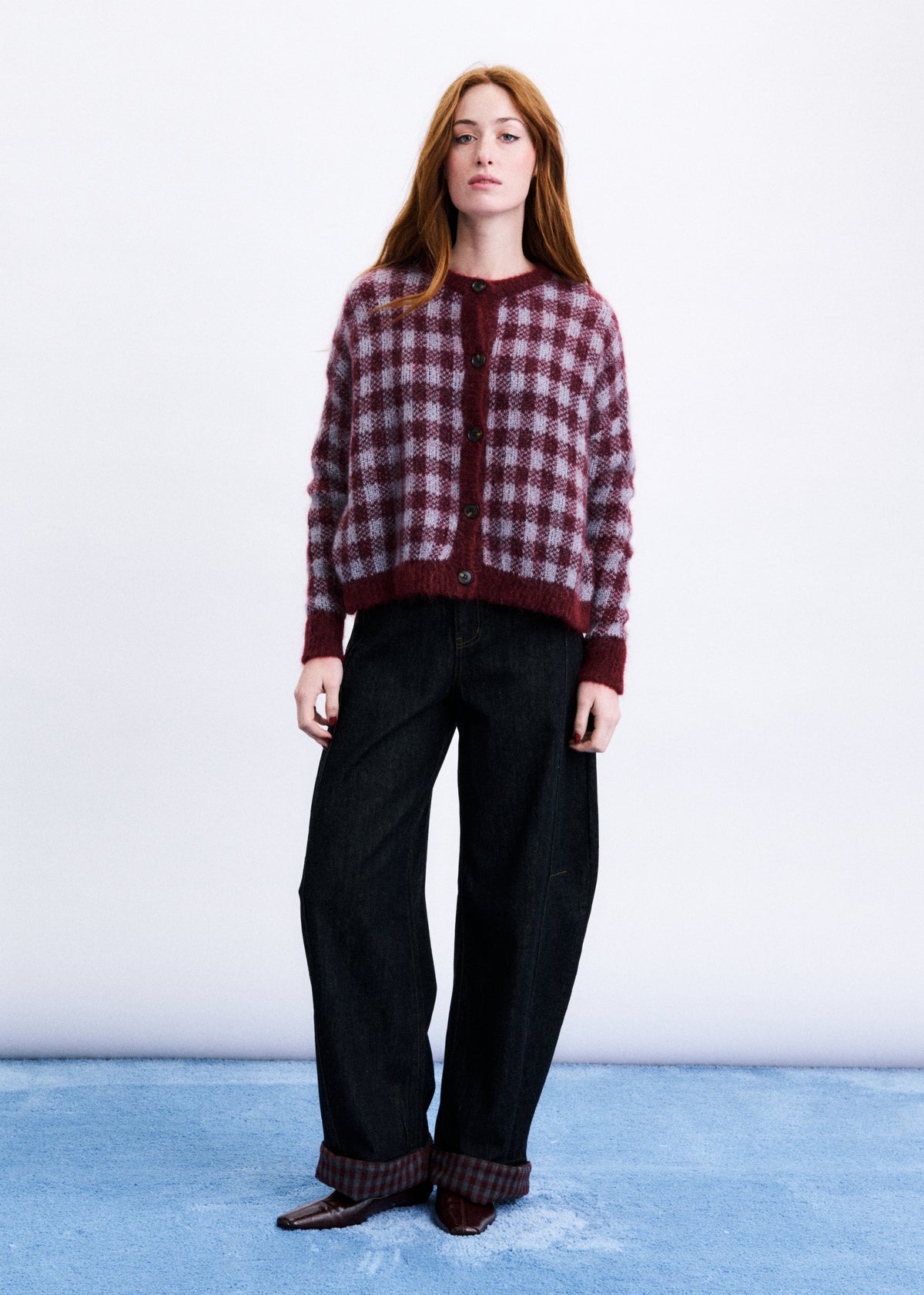 A woman stands against a white studio background wearing a burgundy and dark plaid cardigan with dark wide-leg jeans and brown shoes.
