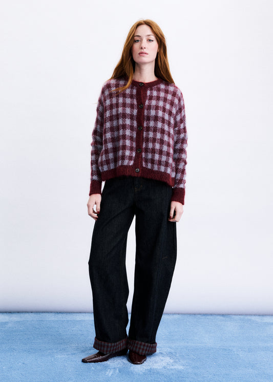 A woman stands against a white studio background wearing a burgundy and dark plaid cardigan with dark wide-leg jeans and brown shoes.