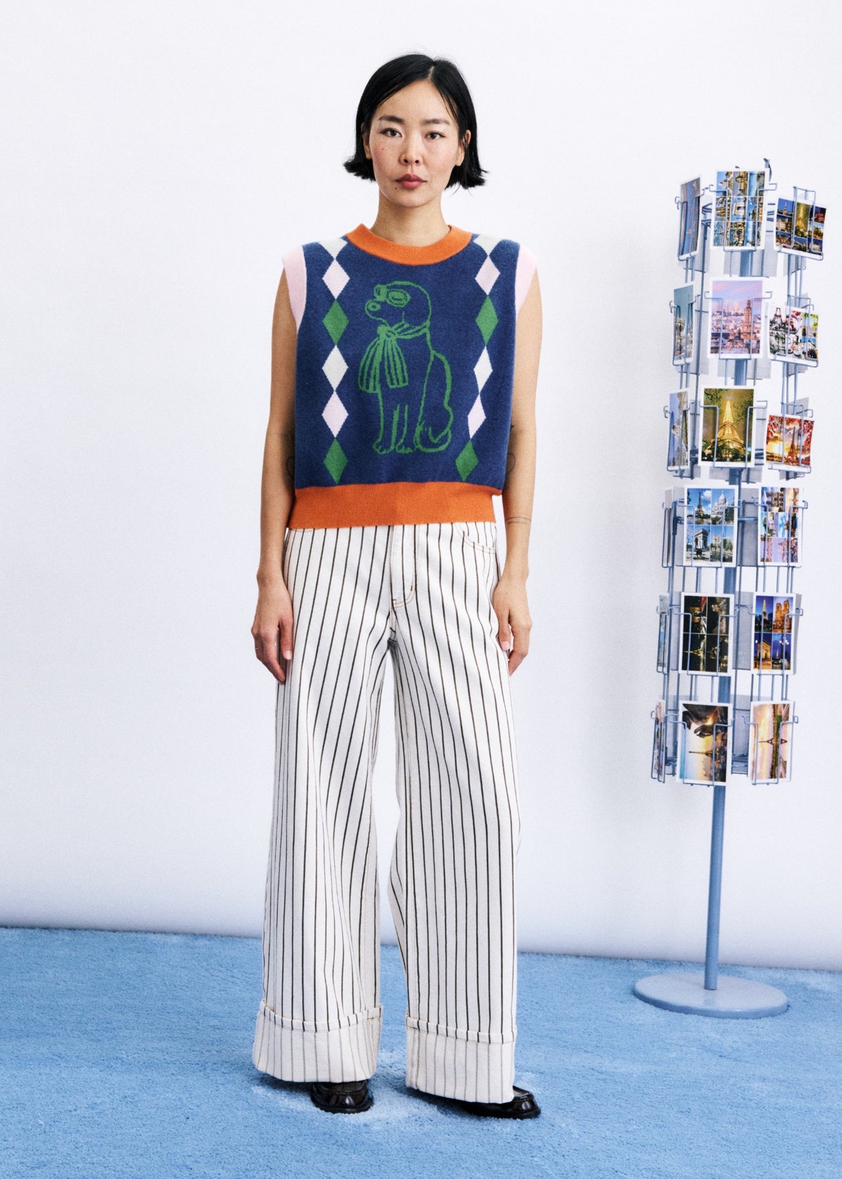 A model stands in a studio wearing a sleeveless knit sweater vest with a green dinosaur graphic and white pinstriped wide-leg trousers. A blue floor and a photo display stand are visible in the background.