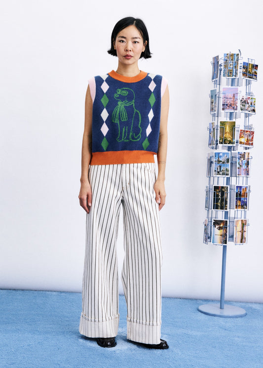 A model stands in a studio wearing a sleeveless knit sweater vest with a green dinosaur graphic and white pinstriped wide-leg trousers. A blue floor and a photo display stand are visible in the background.