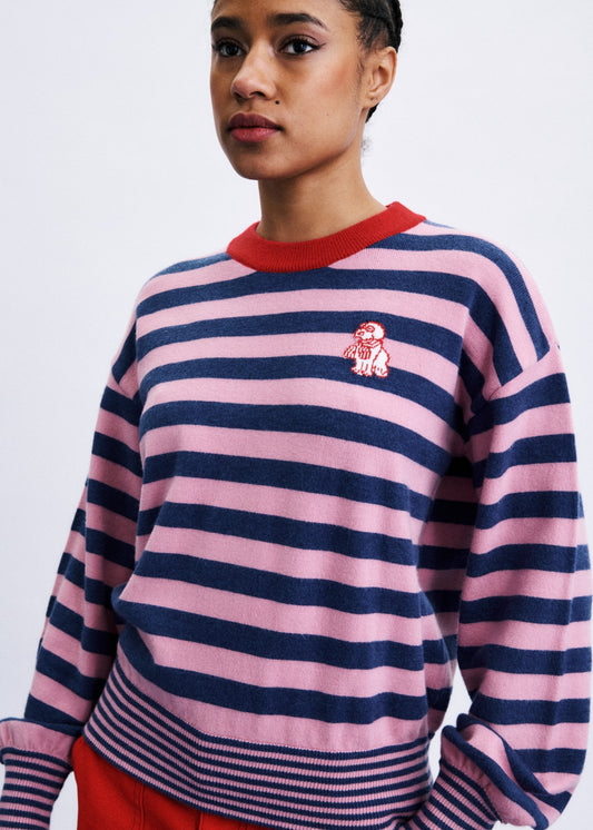 A model wearing a pink and navy blue horizontally striped crewneck sweater with a red collar and a small embroidered character on the left chest, paired with red pants against a light gray background.