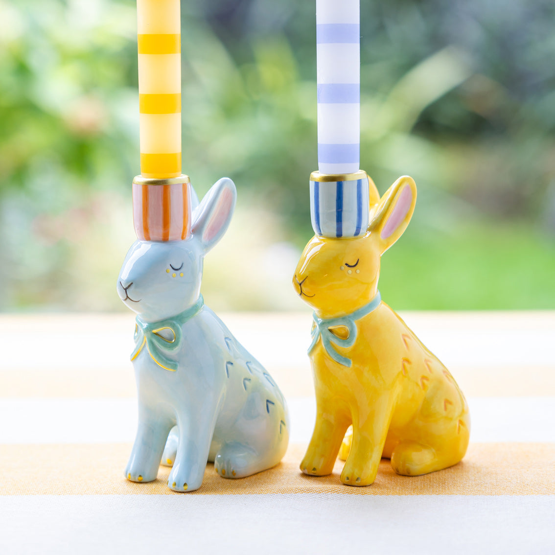 Two ceramic bunny figurines with striped holders on a blurred natural background