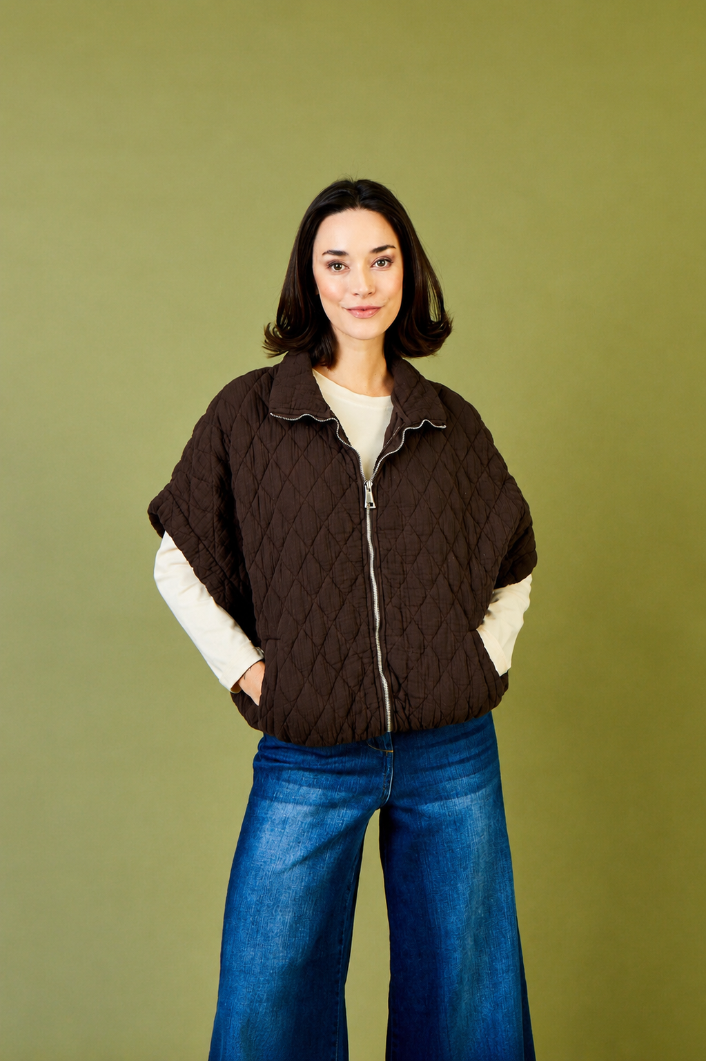 A woman modeling a dark brown diamond-quilted zip-up jacket with white long sleeves, paired with blue jeans, against a solid olive green background.