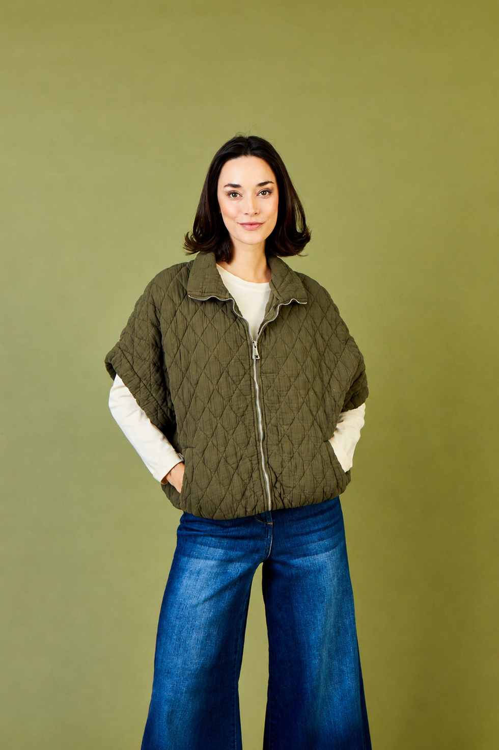 Charlston Quilted Short Sleeve Jacket in Army - a woman modelling a quilted olive green vest over a cream long-sleeve top and blue wide-leg jeans, standing against a solid olive green studio background.