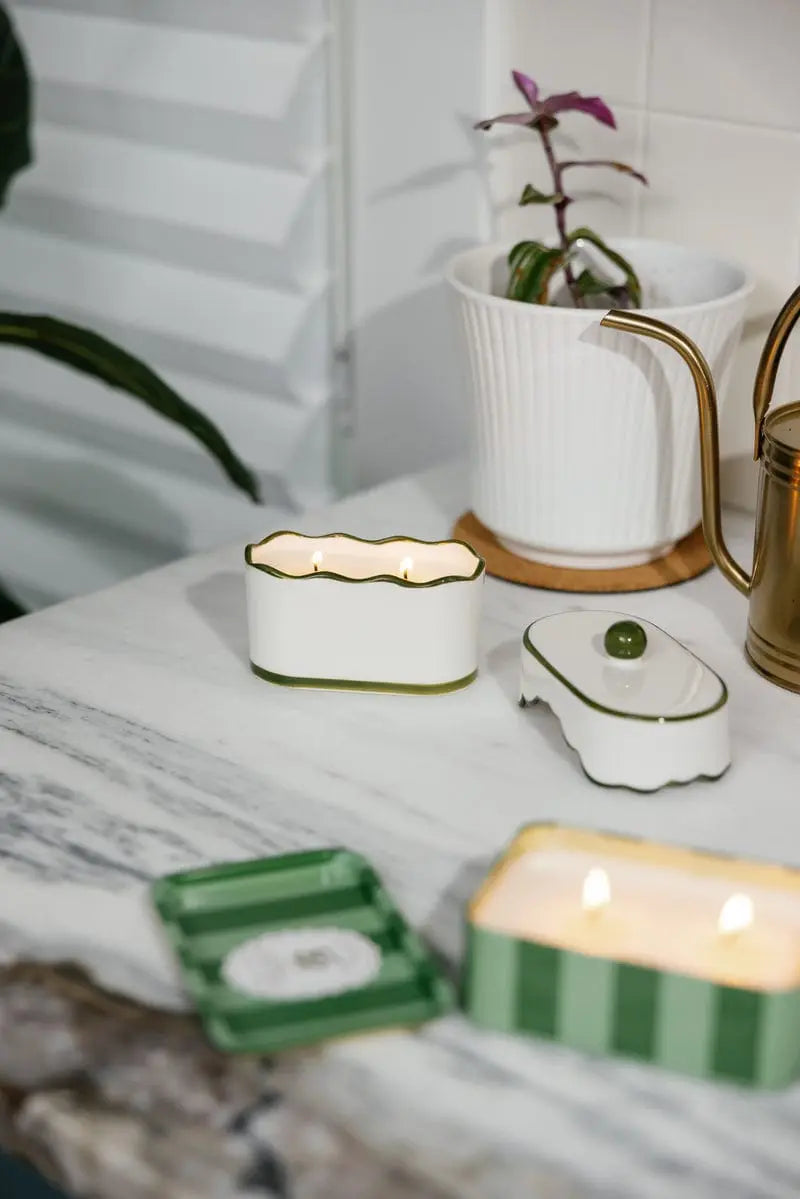 Small white ceramic lanterns with green accents on a surface with a plant and teapot in the background.