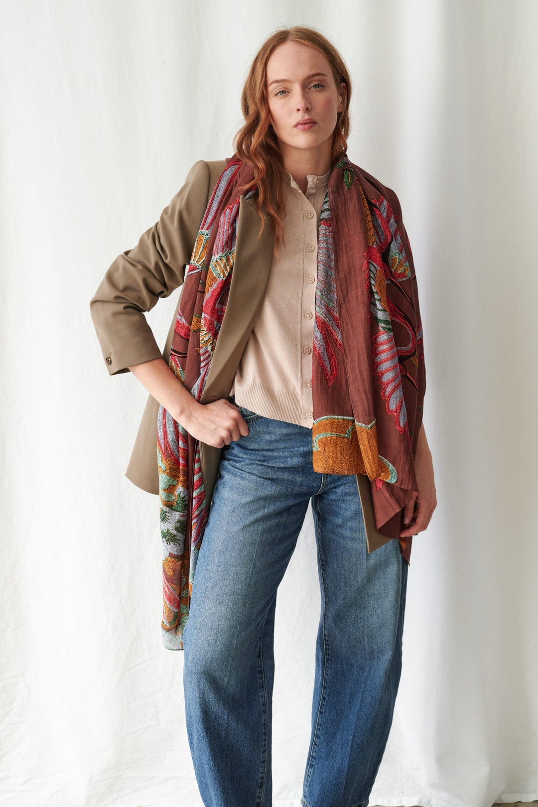 A woman models a long, multicolored patterned scarf draped over her shoulders, styled with a beige blazer, cream shirt, and blue jeans against a white draped fabric background.