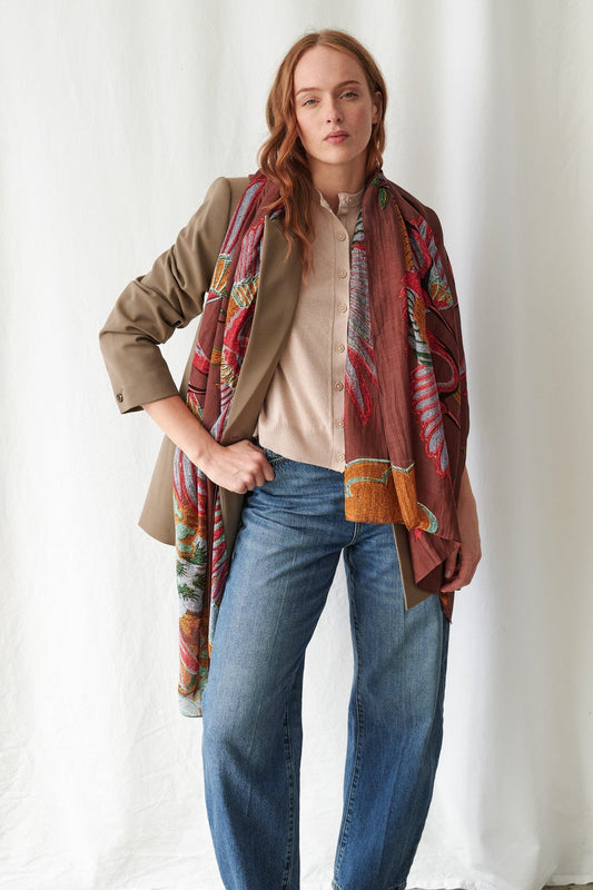 A woman models a long, multicolored patterned scarf draped over her shoulders, styled with a beige blazer, cream shirt, and blue jeans against a white draped fabric background.