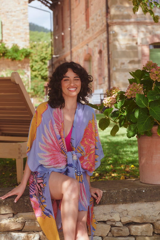 Woman in a colourful outfit sitting outdoors near a stone wall and potted plant.