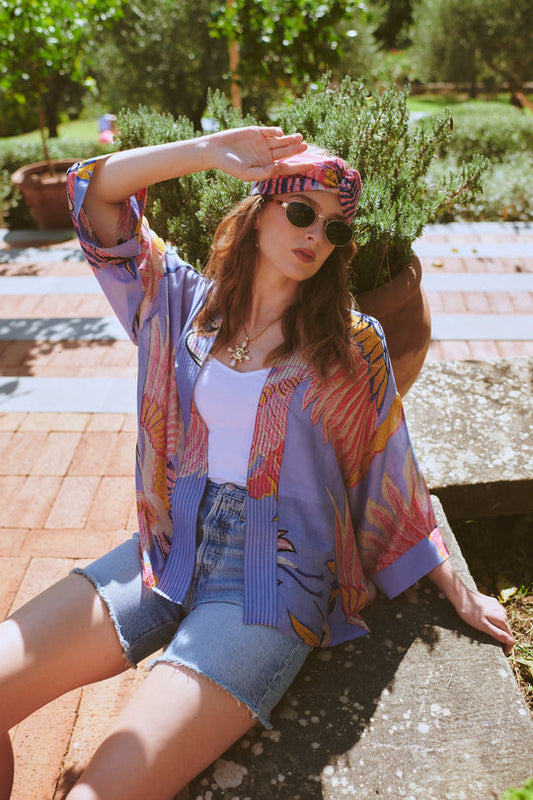 Woman in colourful kimono-style top and denim shorts sitting outdoors with plants in the background