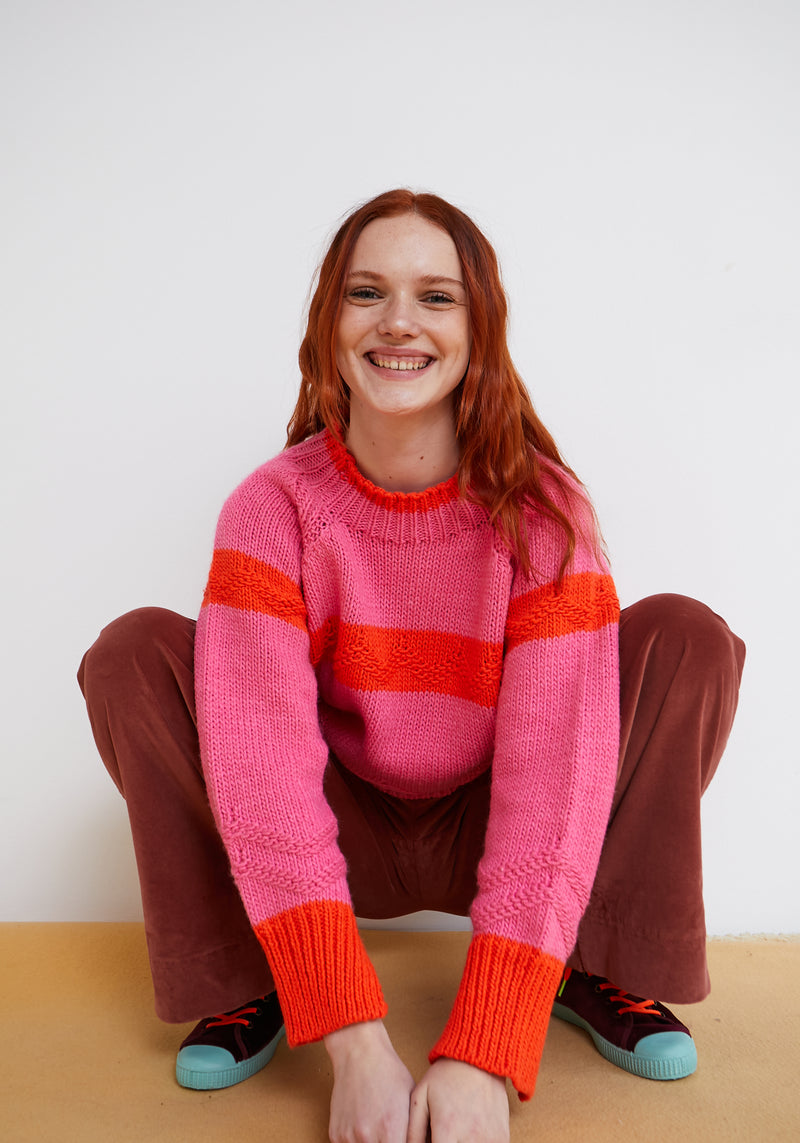 Person wearing a pink sweater with red stripes sitting on a wooden floor.