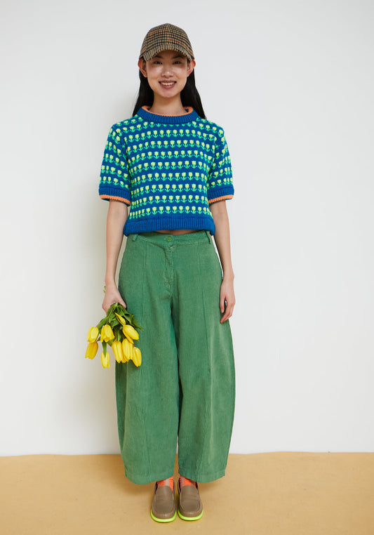 A woman stands against a plain white wall wearing a blue and green patterned short-sleeve knit top, green wide-leg pants, brown shoes, and a brown cap, while holding a small bunch of yellow tulips.