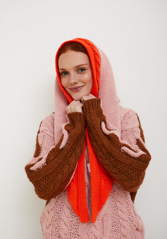 A woman wearing a chunky knit hooded cardigan in pink and brown, with a bright orange scarf wrapped around her neck, posing against a plain white background.