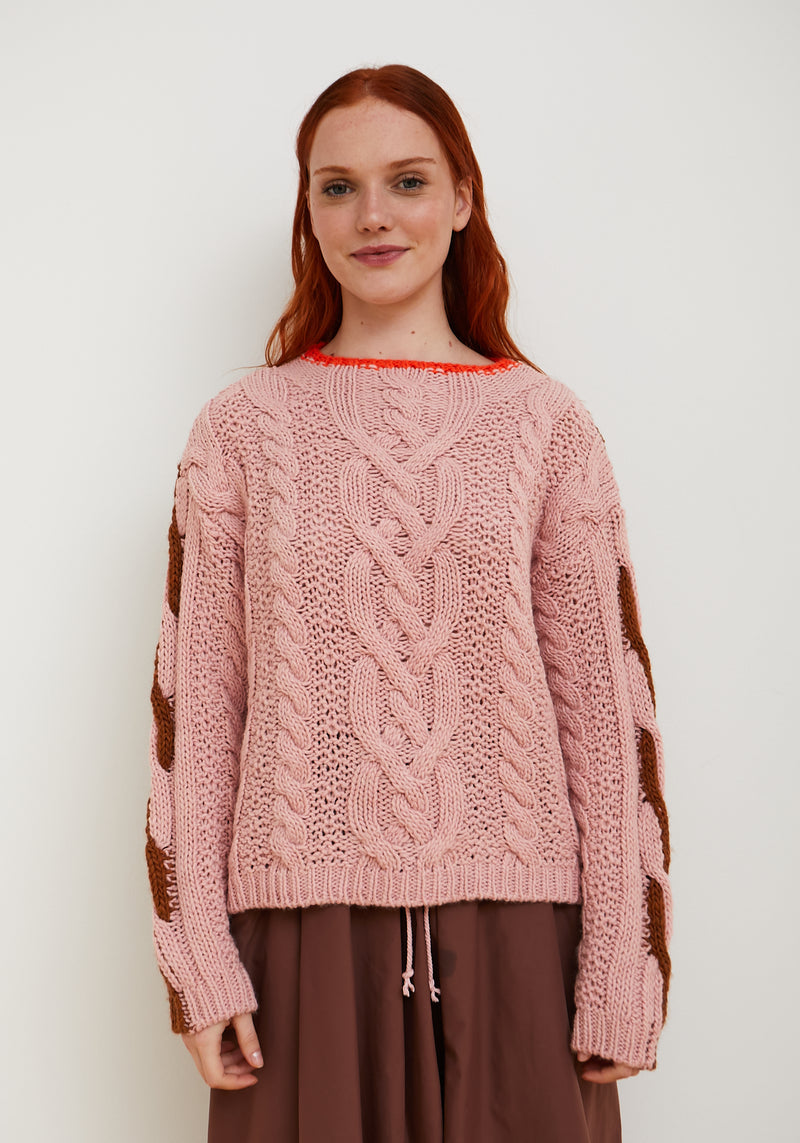 A woman with red hair wearing a pink cable-knit sweater with brown textured knit sleeves, paired with brown pants, standing against a plain white background.