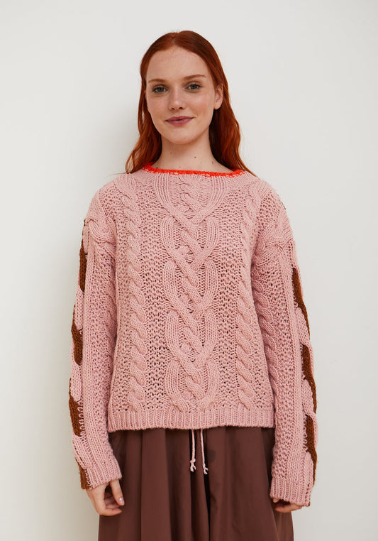 A woman with red hair wearing a pink cable-knit sweater with brown textured knit sleeves, paired with brown pants, standing against a plain white background.