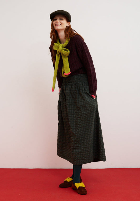 Dr Bloom Cardigan Grace Eggplant. Person wearing a dark coat, green scarf, and plaid skirt on a white background