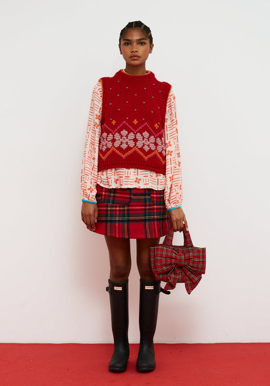 Dr Bloom Jumpy Red Vest. Woman wearing a red patterned sweater, plaid skirt, and black boots on a white background