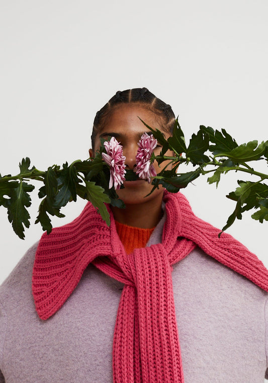 Dr Bloom Spark Pink Collar-Scarf. Person wearing a pink knitted scarf with flowers and leaves in front of their face on a light background