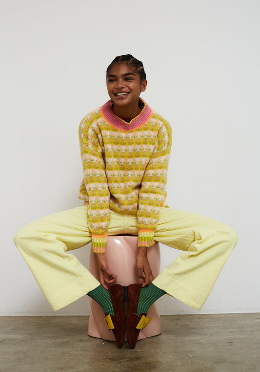 Dr Bloom Sweater Blossom Yellow. Person wearing a colourful sweater and yellow pants sitting on the floor.