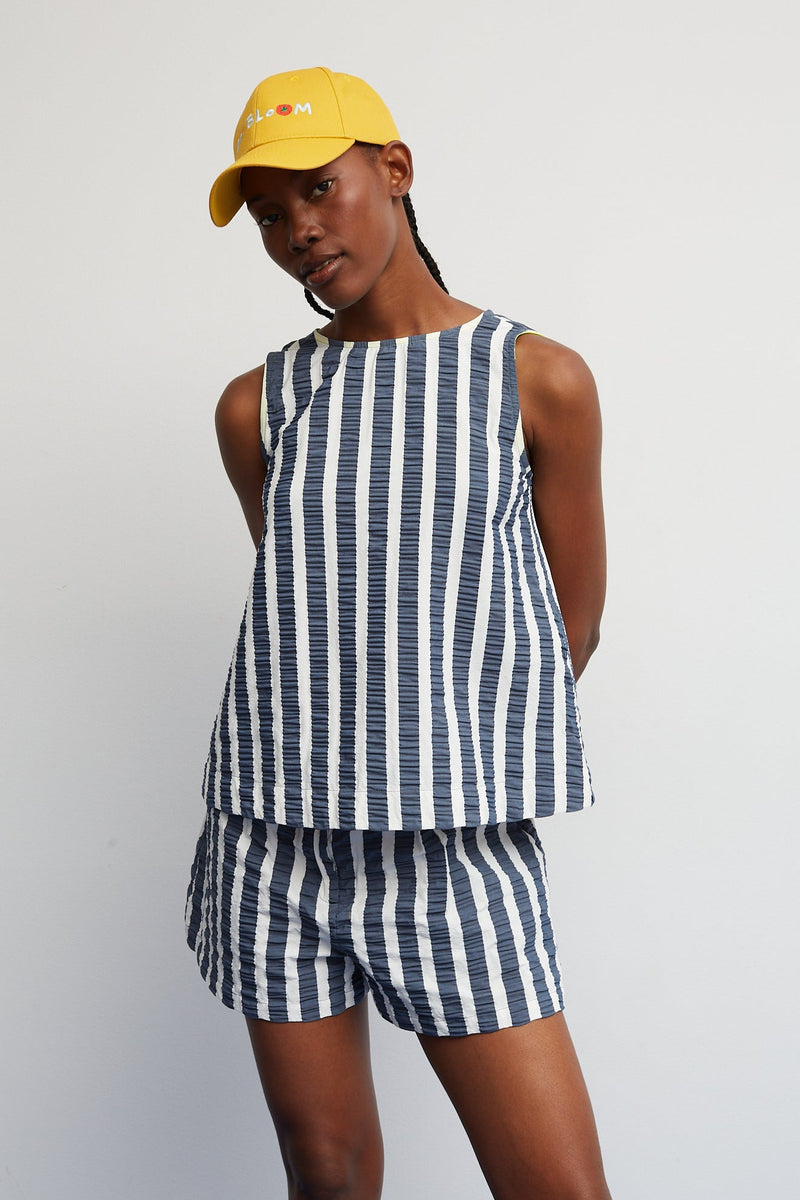 Woman wearing a blue and white striped sleeveless top and shorts with a yellow cap on a white background