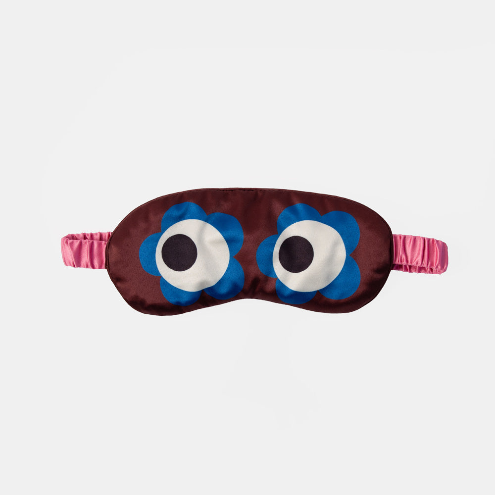 A sleep mask with a dark brown satin body, featuring two large blue flower-shaped eyes with white and black centers, and a pink elastic strap, shown against a light gray background.