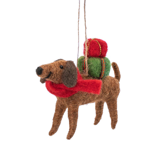 A felted wool dog Christmas ornament in brown with a red scarf, carrying a stack of red and green wrapped gifts on its back, hanging from a string against a white background.