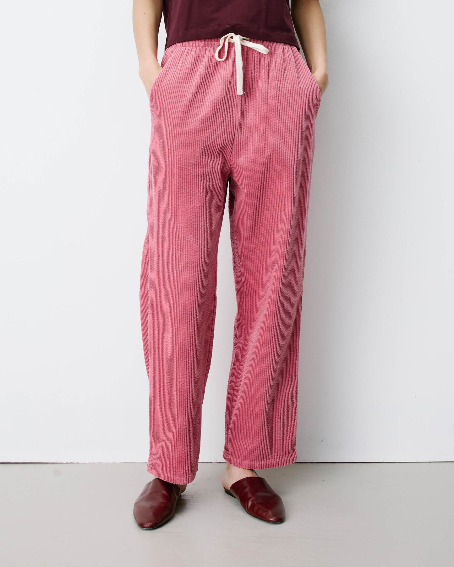 Grace & Mila Liberty Trouser in Blush 
A person wearing loose, wide-leg pink textured lounge pants with a cream drawstring waist, paired with a dark burgundy top and burgundy loafers, standing against a plain light gray wall.
