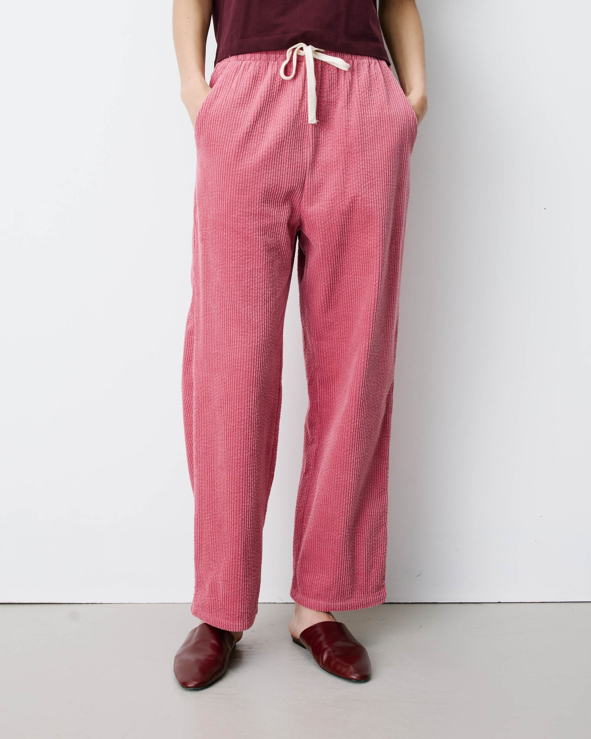 Grace & Mila Liberty Trouser in Blush 
A person wearing loose, wide-leg pink textured lounge pants with a cream drawstring waist, paired with a dark burgundy top and burgundy loafers, standing against a plain light gray wall.
