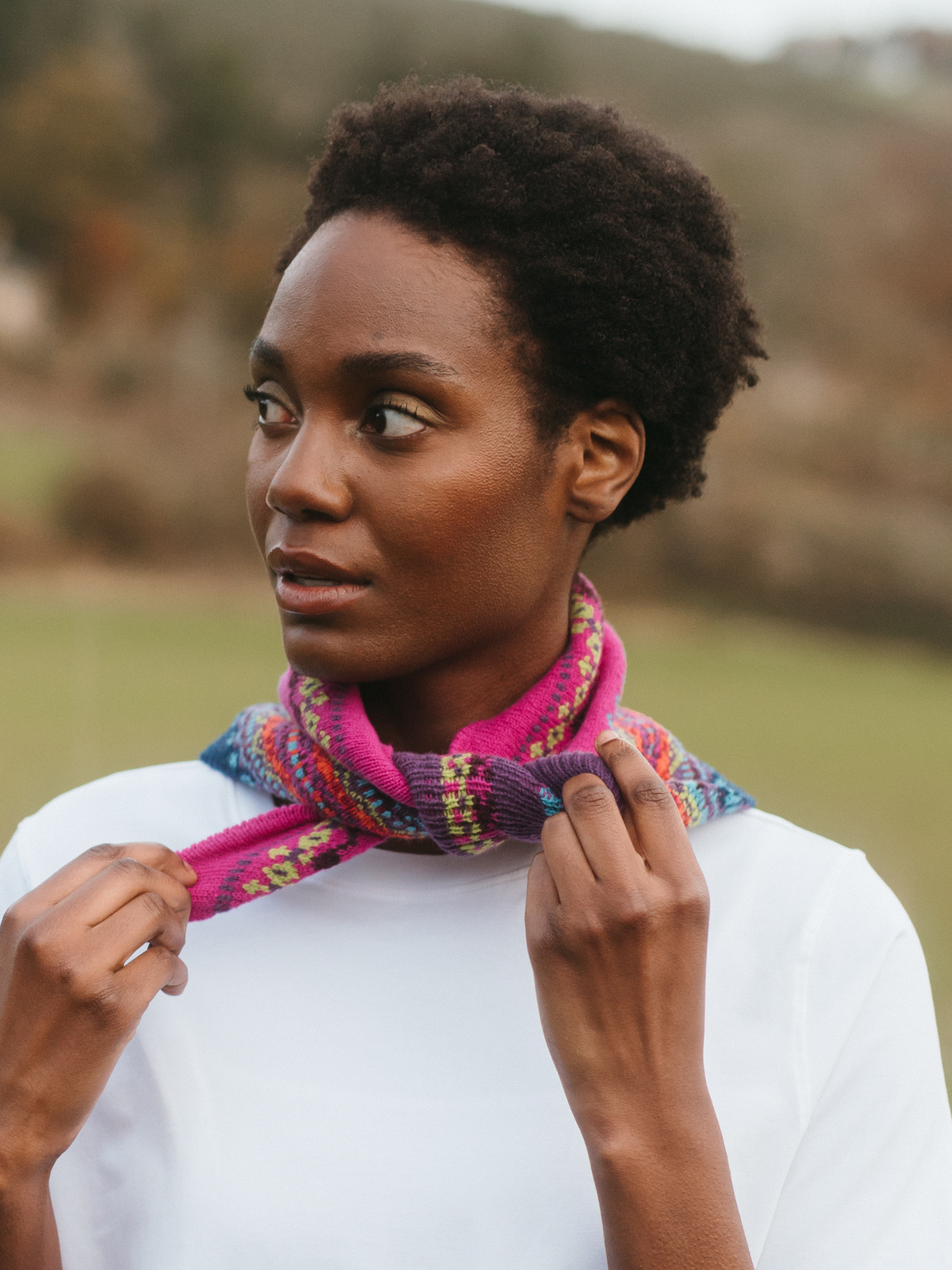 A woman wearing a vibrant pink and multicolor patterned knit scarf tied around her neck, paired with a white t-shirt, set against a soft-focus outdoor background.