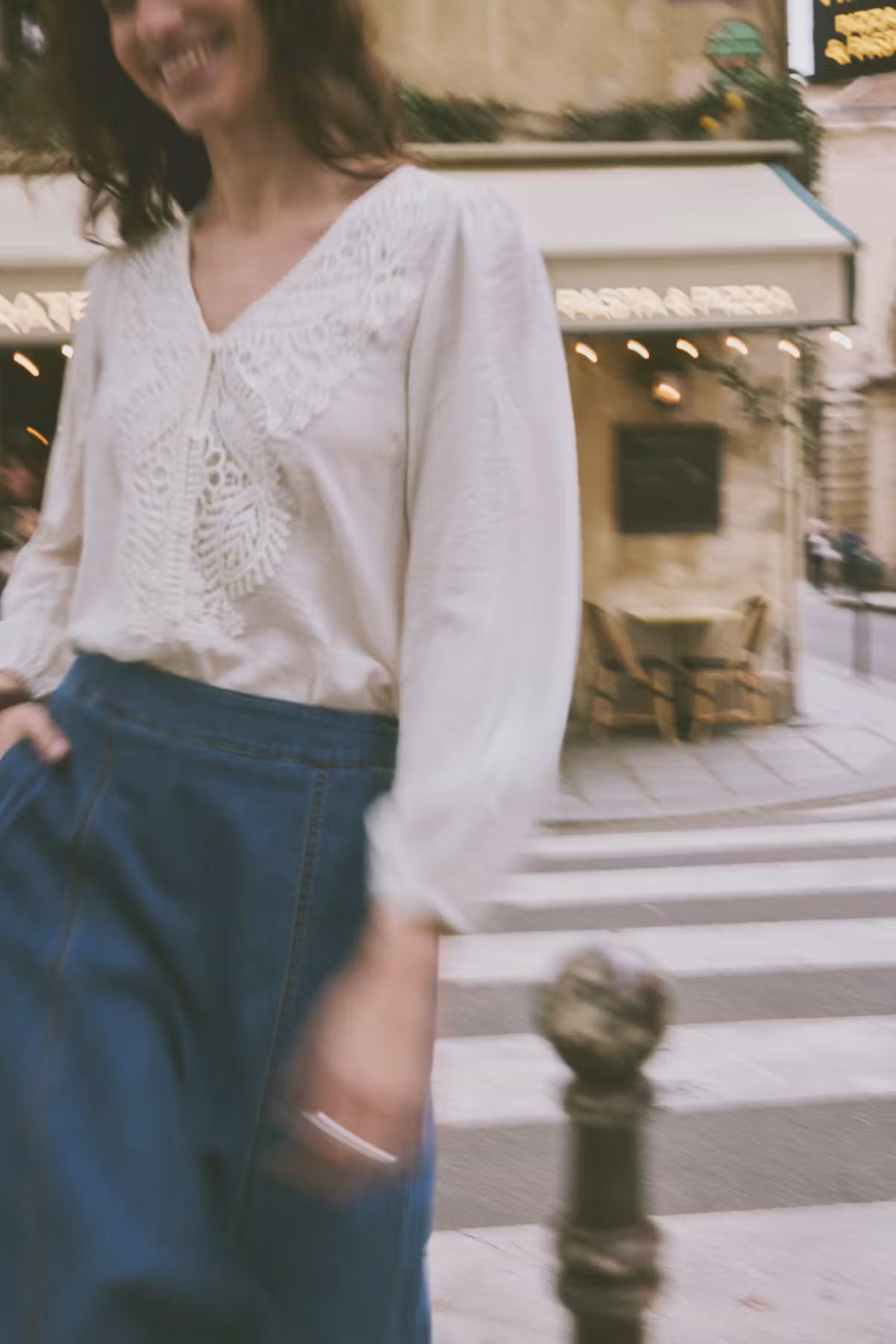 A woman wearing a white long-sleeve blouse with delicate lace embroidery on the chest and high-waisted dark blue trousers, standing on a city street crosswalk in front of a cafe.