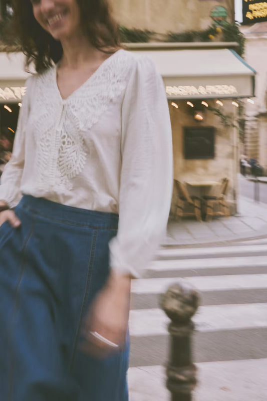 A woman wearing a white long-sleeve blouse with delicate lace embroidery on the chest and high-waisted dark blue trousers, standing on a city street crosswalk in front of a cafe.