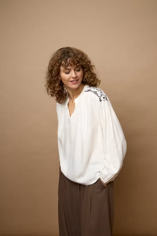 A woman with curly brown hair wearing a white long-sleeve blouse with black floral embroidery on the shoulder and dark brown wide-leg trousers, posing against a solid warm brown studio backdrop.