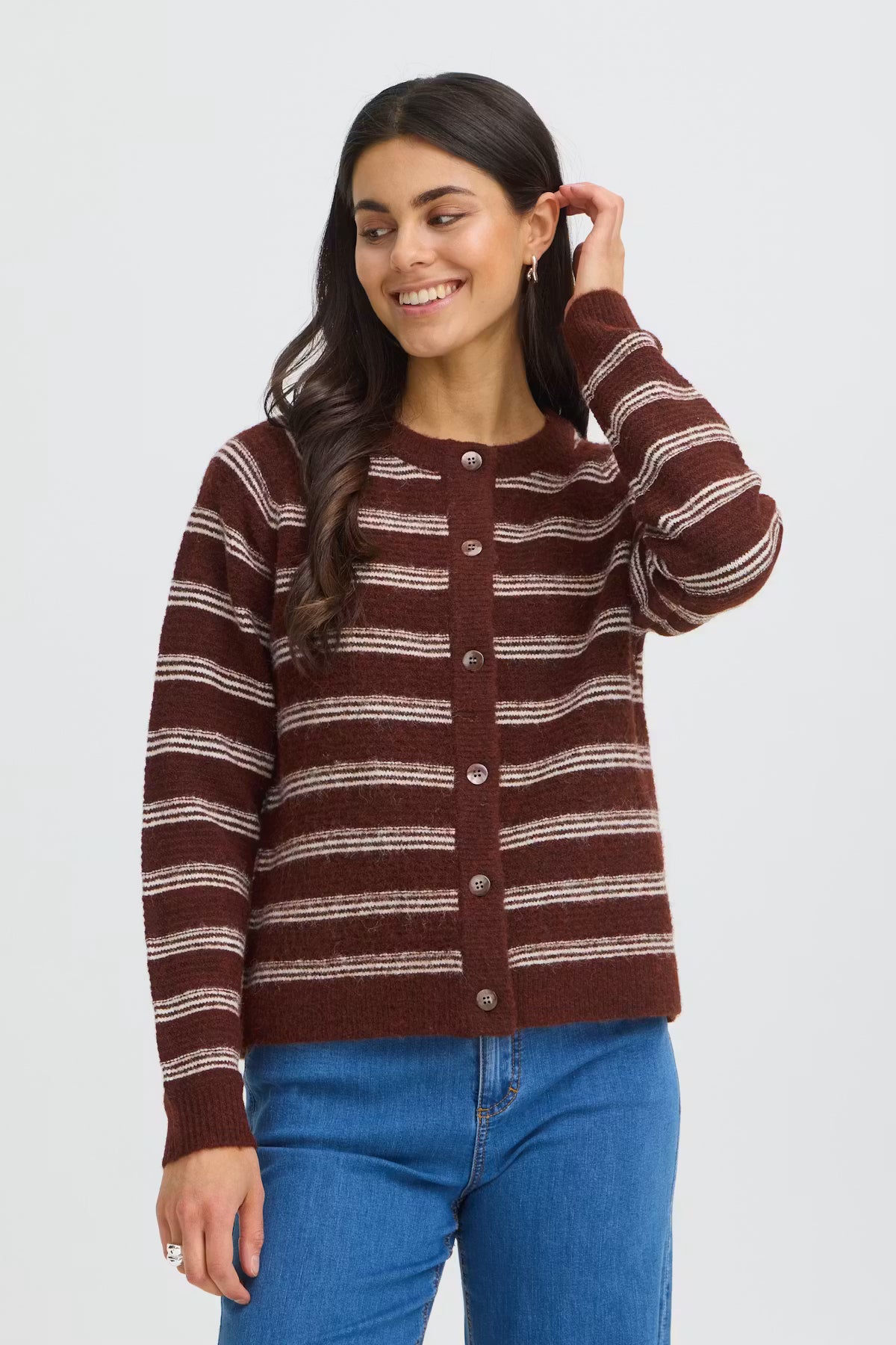 A woman wearing a rusty brown and beige horizontally striped knit cardigan with a button-up front, paired with blue jeans, against a light gray studio background.