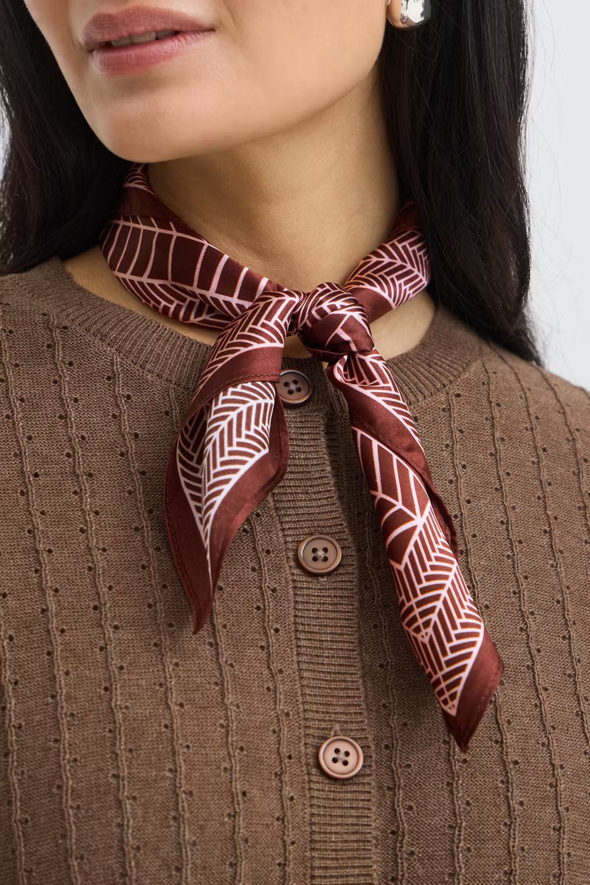 A close-up of a woman wearing a brown ribbed knit cardigan sweater and a patterned silk neck scarf tied around her collar. The scarf features a geometric chevron design in dark red and white.