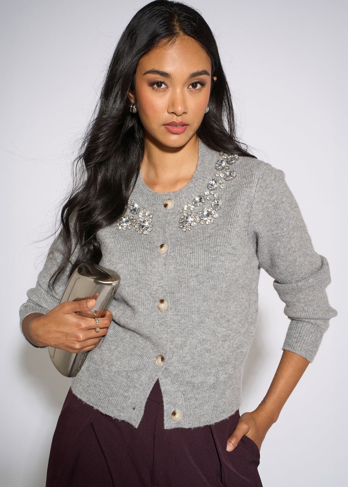 A woman modeling a light gray knit cardigan with crystal embellishments around the neckline, paired with dark plum pants and holding a metallic clutch against a plain light gray background.