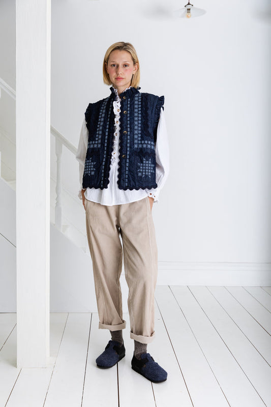 Person wearing a dark blue patterned vest over a white shirt with beige pants on a white wooden floor.