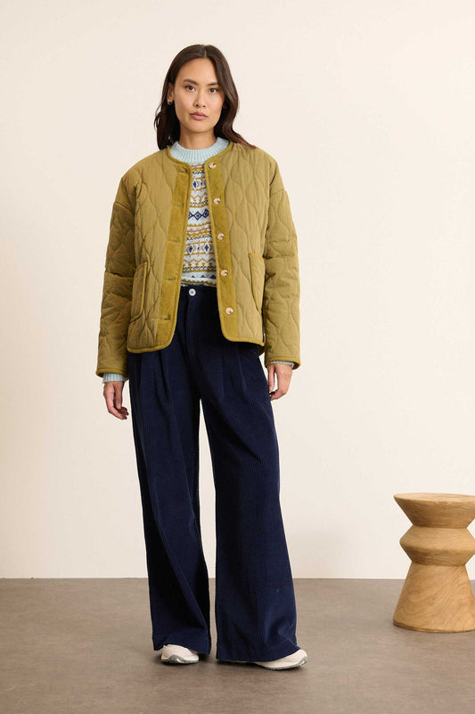 Woman wearing a green jacket and navy pants standing against a beige wall.