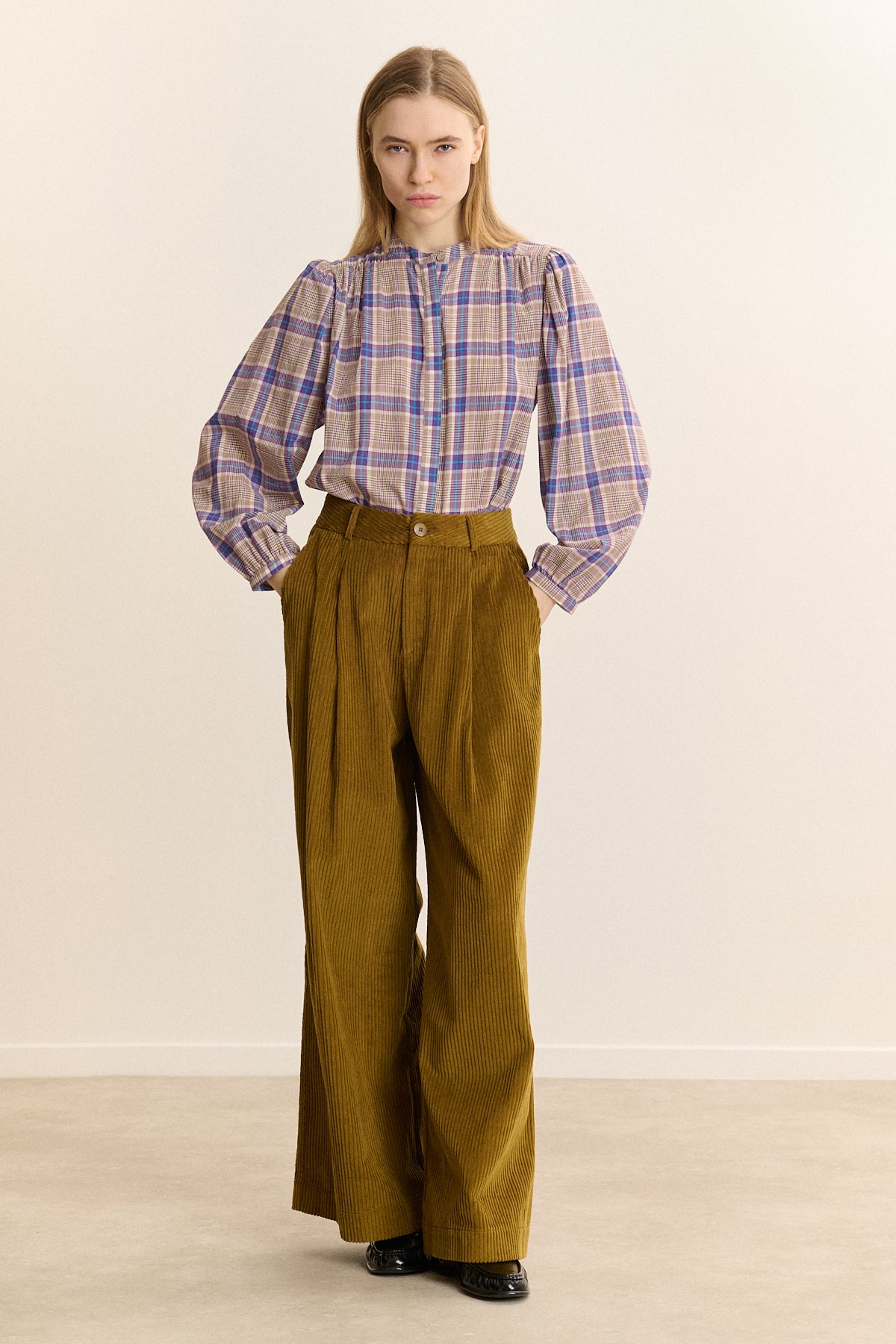 A woman stands in a studio wearing a long-sleeve plaid button-up shirt in beige, purple, and blue, paired with high-waisted olive-brown wide-leg trousers and black shoes.
