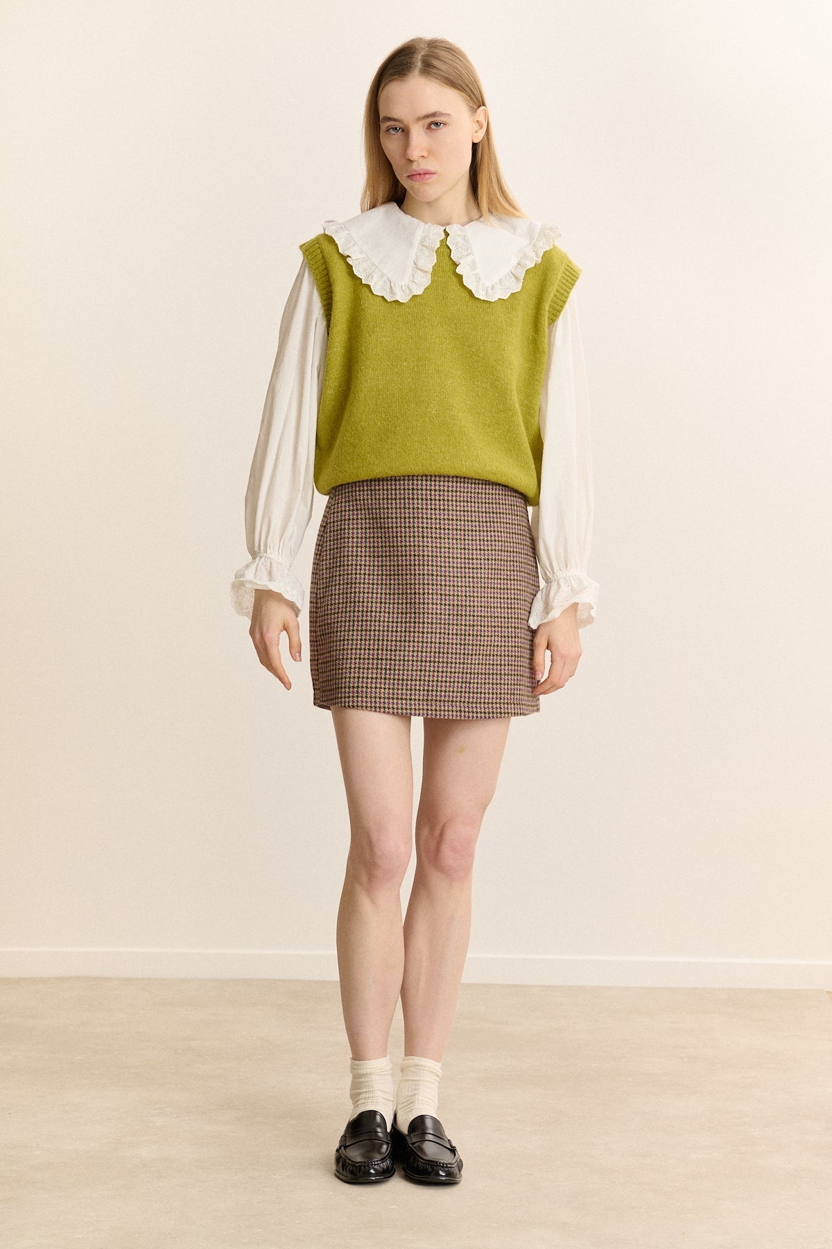 Garance Paris Jewel Sweater in Olive - a model stands against a plain beige studio wall wearing a sleeveless olive green knit sweater vest over a white long-sleeve blouse with a large ruffled collar, paired with a brown and beige houndstooth mini skirt, white socks, and black loafers.