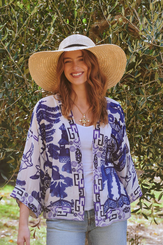 Woman wearing a blue and white patterned kimono with a wide-brimmed hat in front of green foliage.