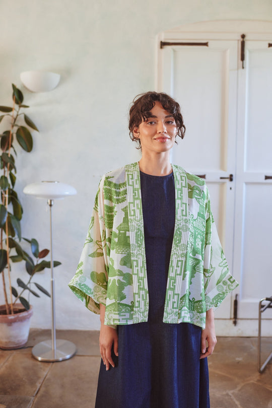 Woman wearing a green patterned kimono over a dark dress in a room with plants and a lamp.