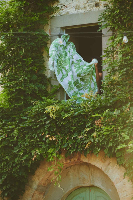 Person holding a green and white patterned fabric out of an ivy-covered window.