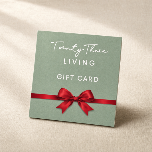A square mint green gift card with white text reading 'Twenty Three LIVING GIFT CARD' and a red ribbon bow across the lower middle, photographed on a beige fabric background.