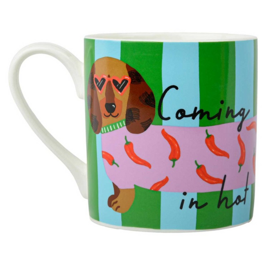 A white ceramic mug featuring a whimsical dachshund illustration wearing heart-shaped sunglasses and a pink sweater with red chili peppers, set against green and light blue vertical stripes. The text 'Coming in hot' is written in black script on the right side.