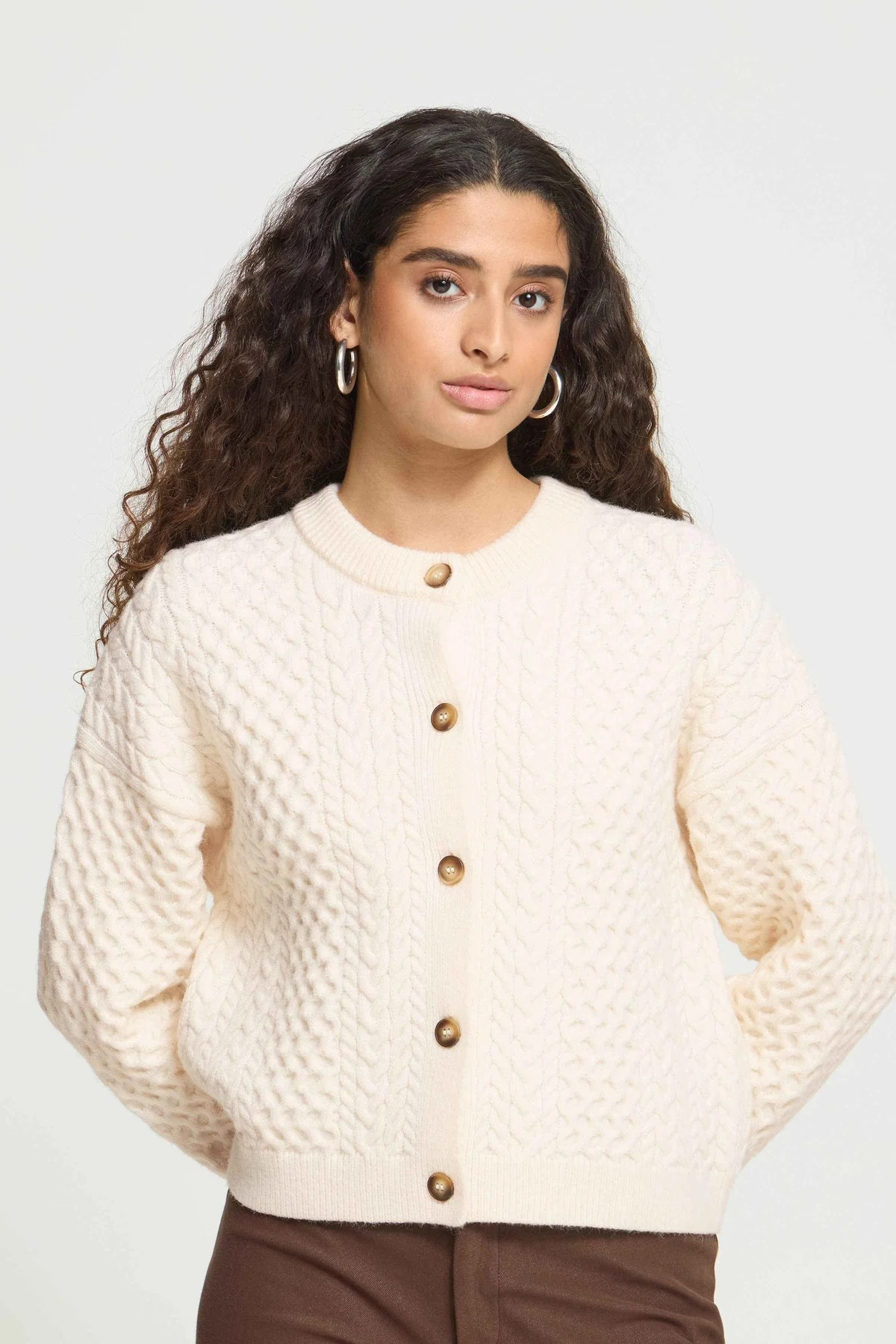 A woman with long, curly dark brown hair wearing a cream-colored cable-knit cardigan with brown buttons, paired with brown pants, against a plain light gray studio background.