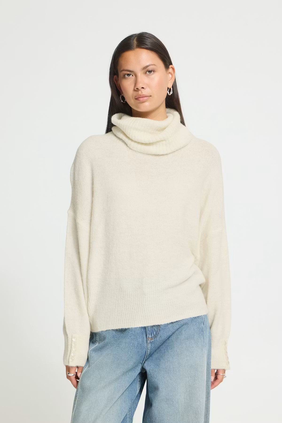A woman modeling a cream-coloured knit cowl neck sweater with light blue jeans, photographed against a solid light grey background.