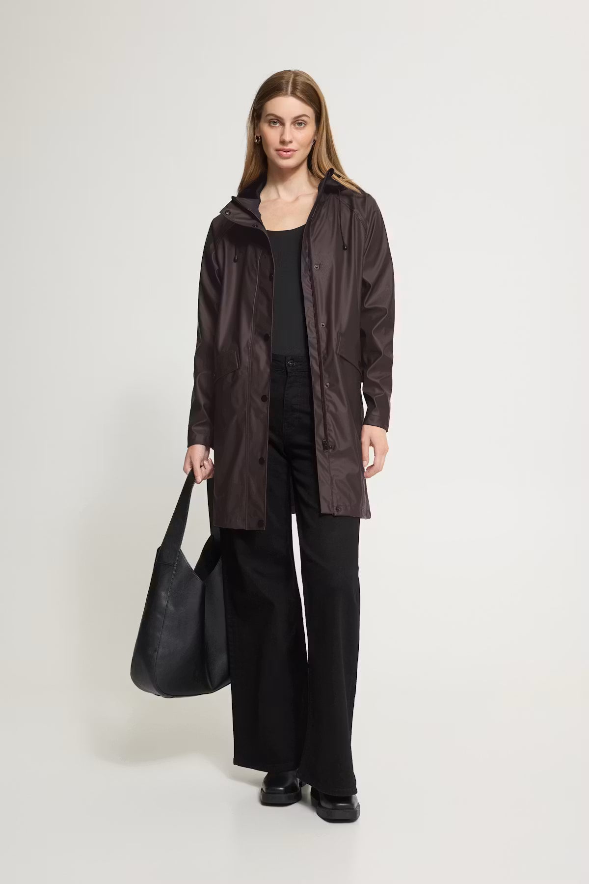 A woman stands against a light gray studio background wearing a dark brown hooded rain jacket over a black top and black wide-leg pants, while holding a large black tote bag.