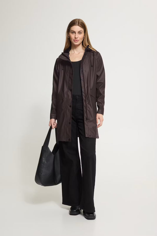 A woman stands against a light gray studio background wearing a dark brown hooded rain jacket over a black top and black wide-leg pants, while holding a large black tote bag.