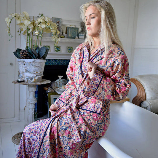 A woman wearing a pink and blue floral print robe sits on a white bathtub in a bright, white-themed bedroom.