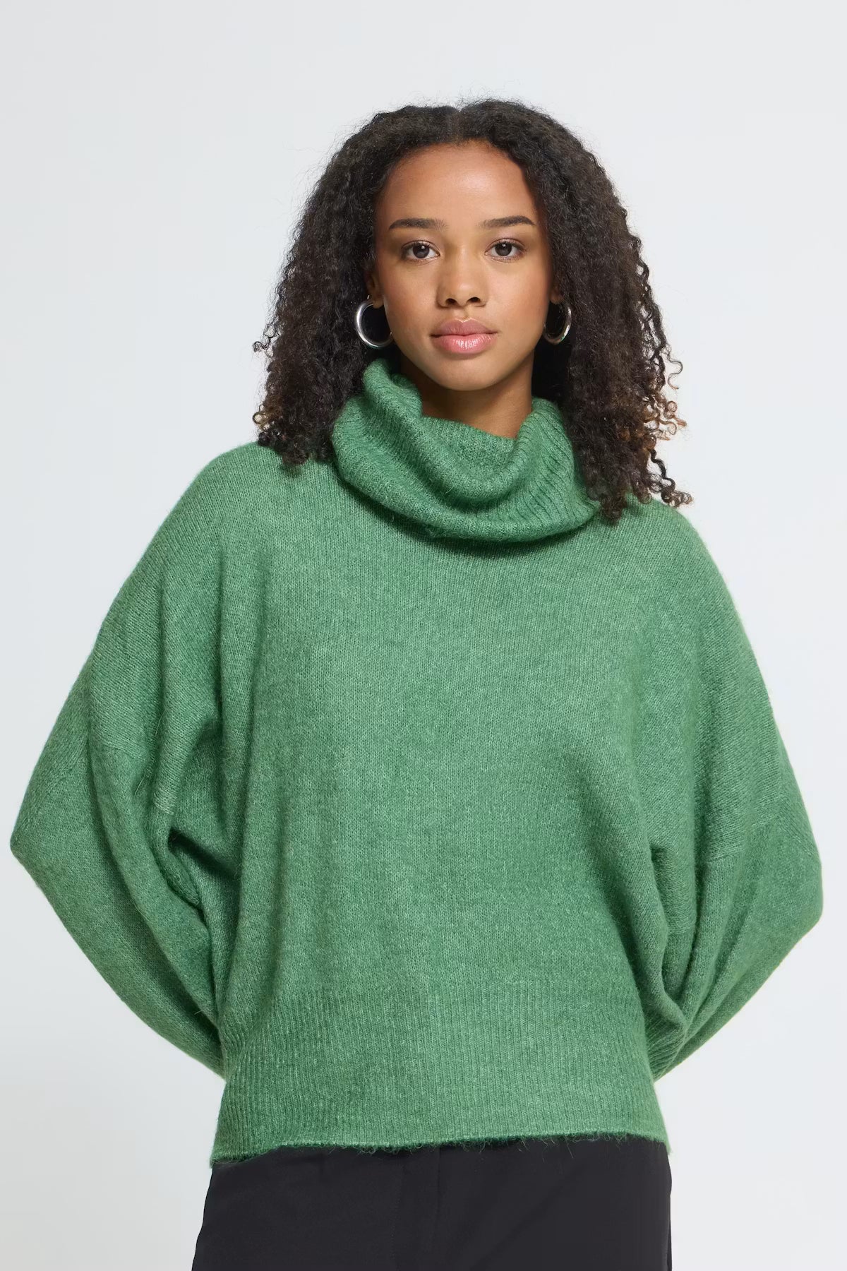 A woman wearing a green knit turtleneck sweater with wide batwing sleeves, paired with black pants, against a plain light gray studio background.