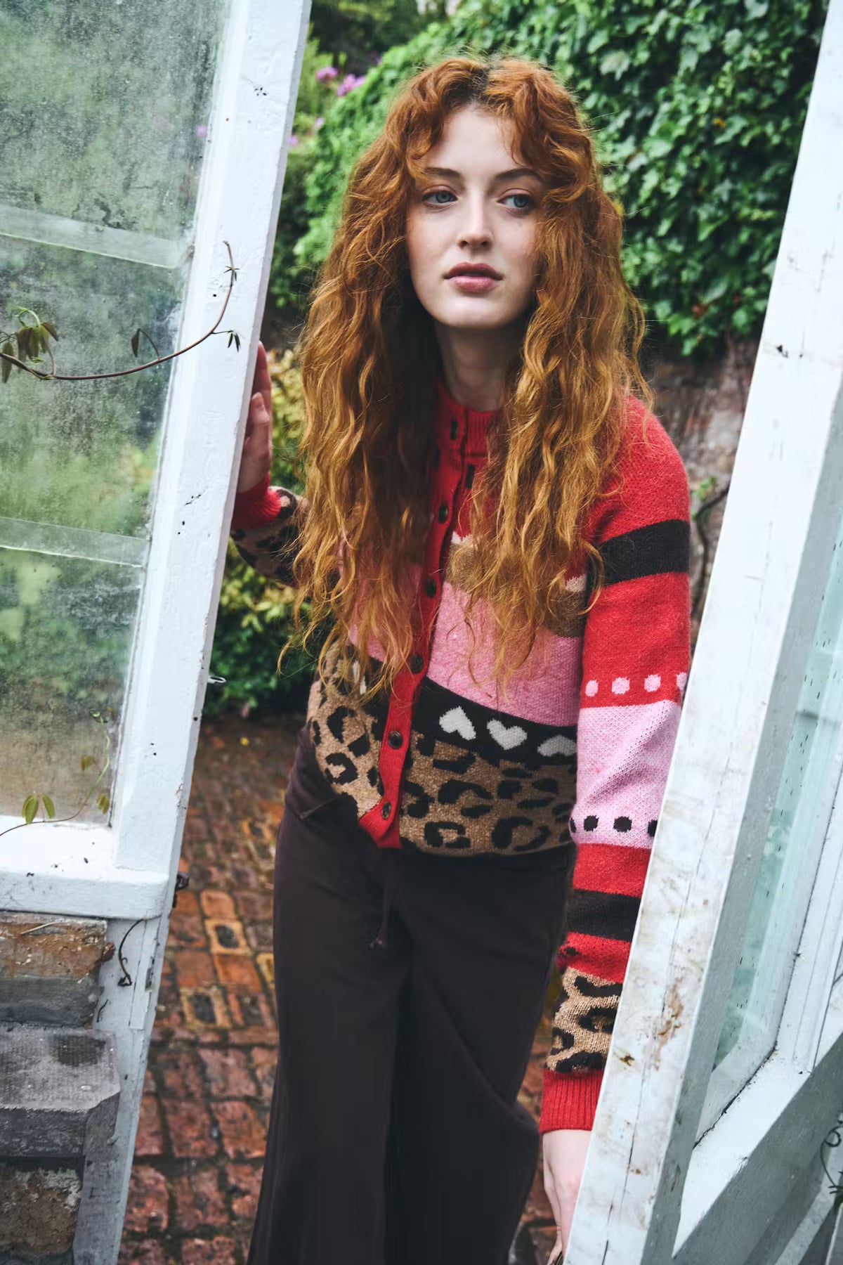 A woman with long, wavy red hair stands in an open white greenhouse doorway, wearing a color-blocked knit cardigan with leopard print, pink, red, and black panels, paired with dark brown pants.